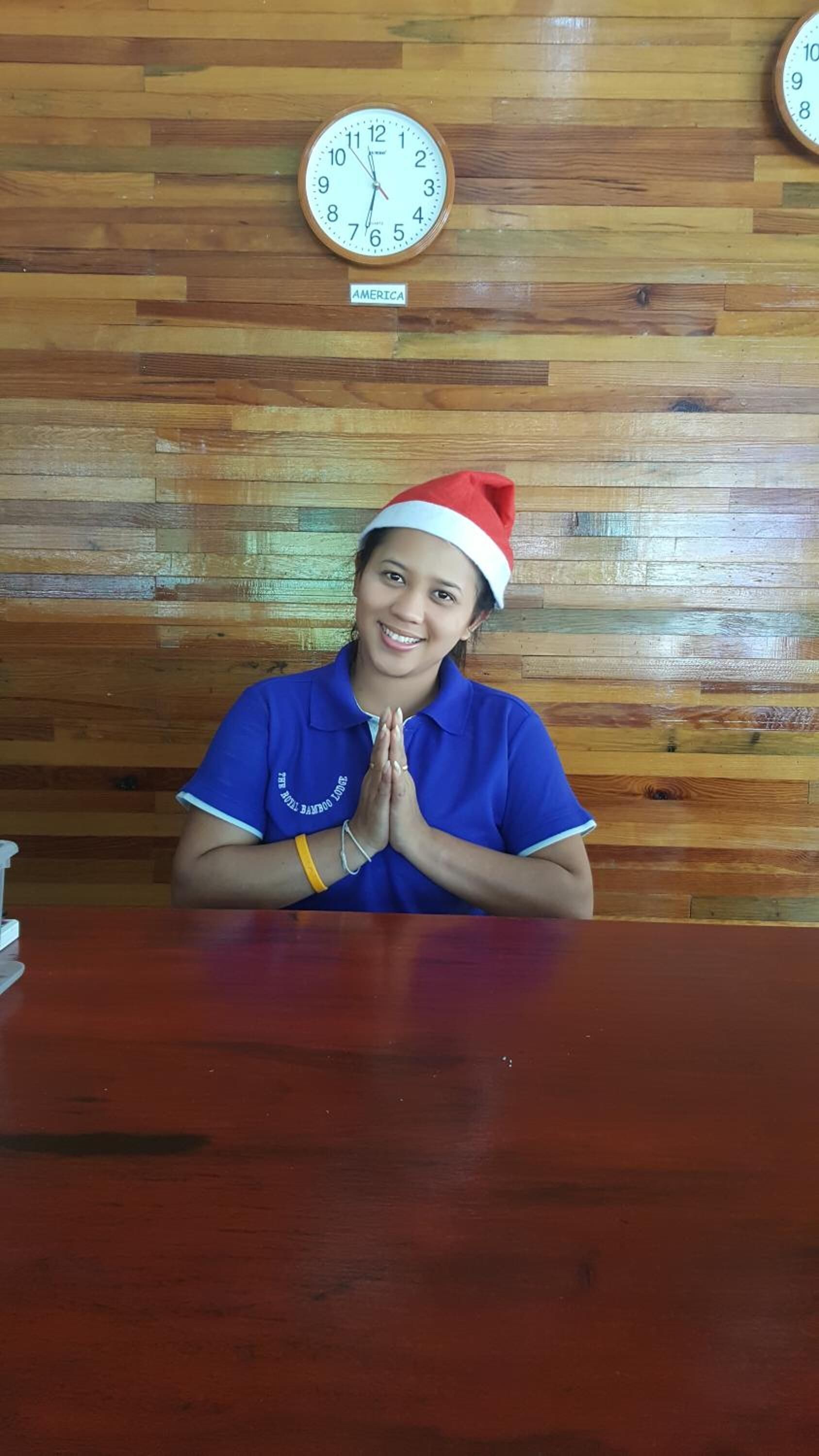 Una persona con una camicia blu e un cappello da Babbo Natale seduta a un tavolo di legno con un orologio appeso al muro sullo sfondo.
