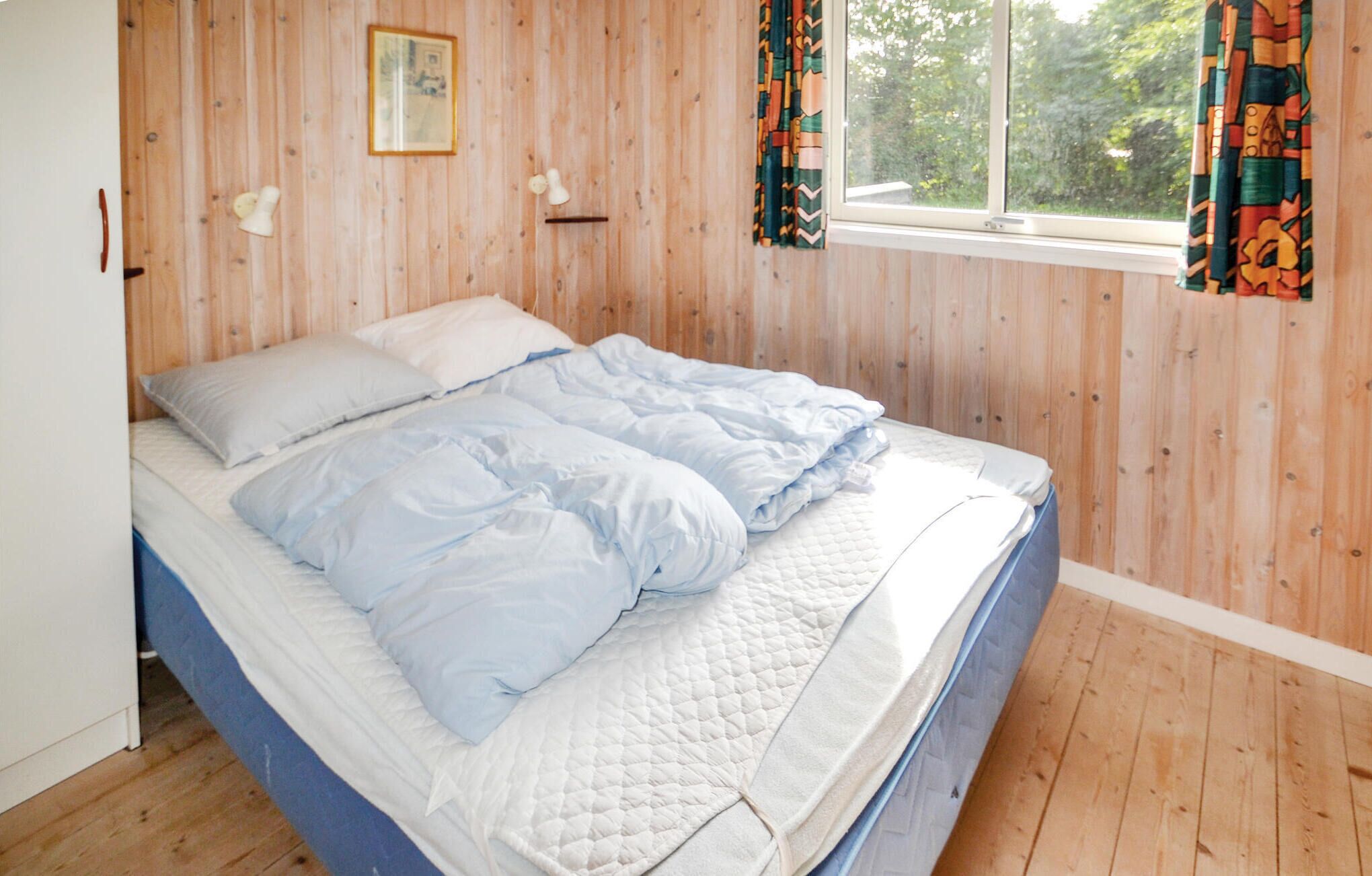 A bedroom with a wooden floor, a bed with white and blue bedding, a window with curtains, and a picture on the wall.
