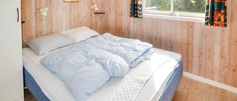 A bedroom with a wooden floor, a bed with white and blue bedding, a window with curtains, and a picture on the wall.