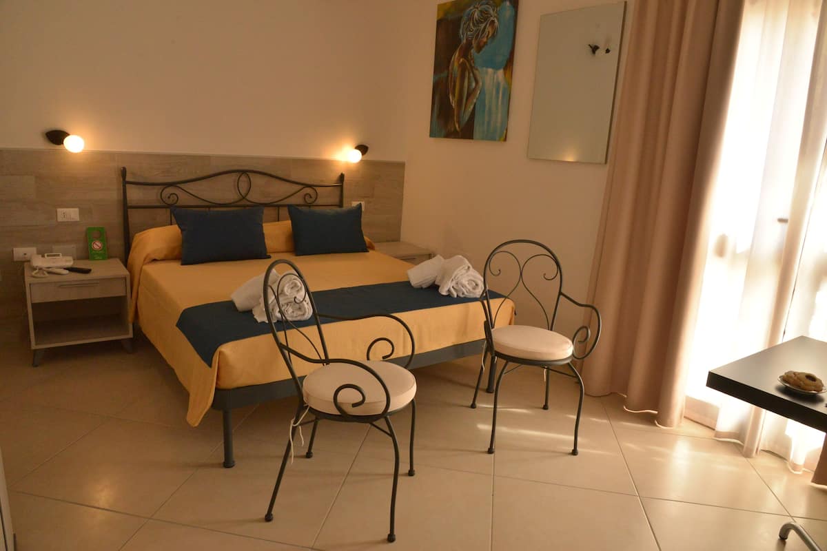 A hotel room with a bed, two chairs, a nightstand, a table with a plate, and a painting on the wall.