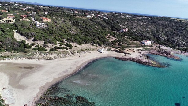An aerial view of a coastal area with a sandy beach, clear turquoise waters, and scattered buildings on the hillside. - Residenza Al Castello (Carloforte)