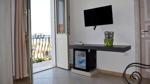 A room with a sliding glass door leading to a balcony, a mounted TV, a small black refrigerator, and a shelf with decorative items. - Residenza Al Castello (Carloforte)
