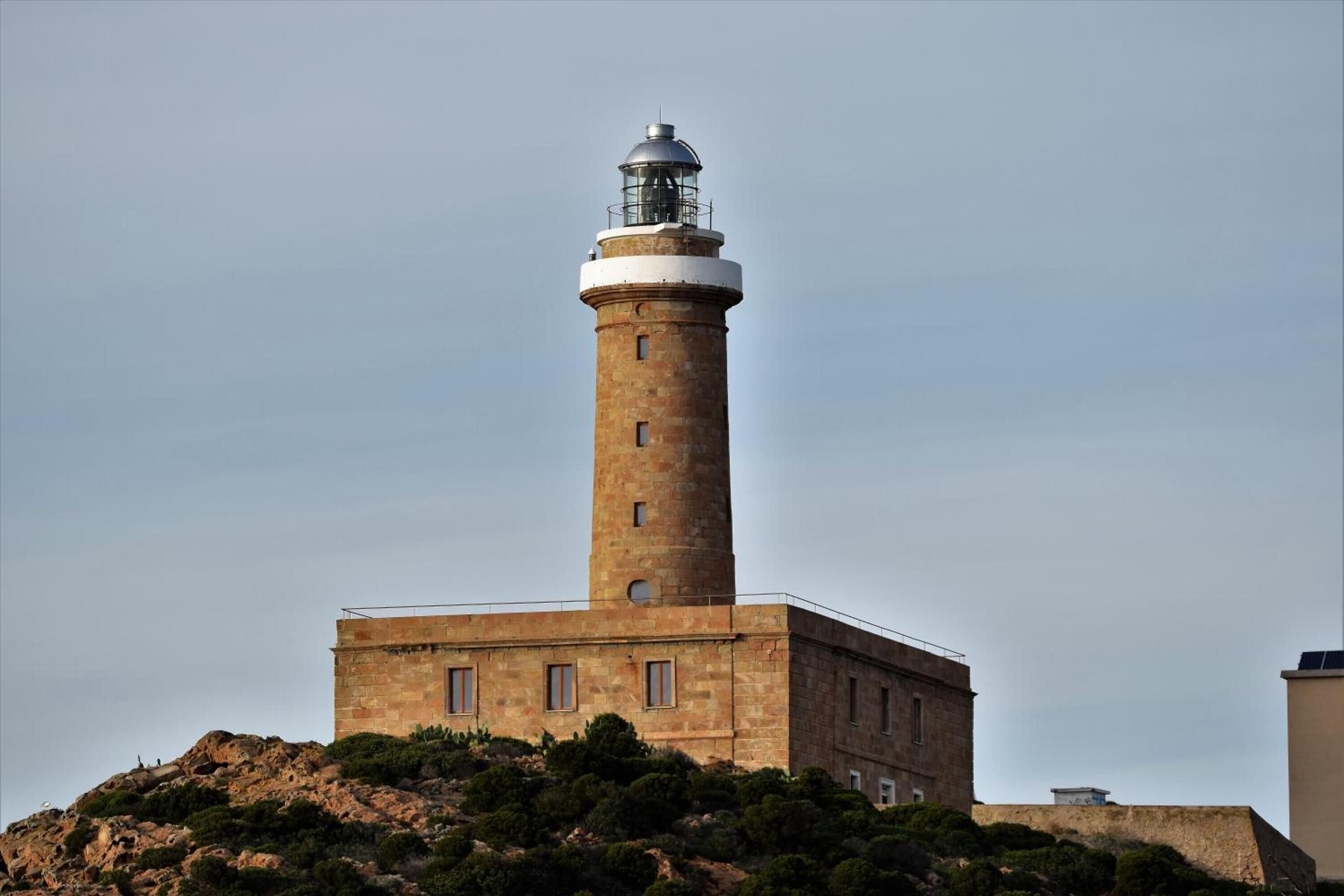 A lighthouse with a cylindrical tower and a flat base, situated on a rocky hill with sparse vegetation.
