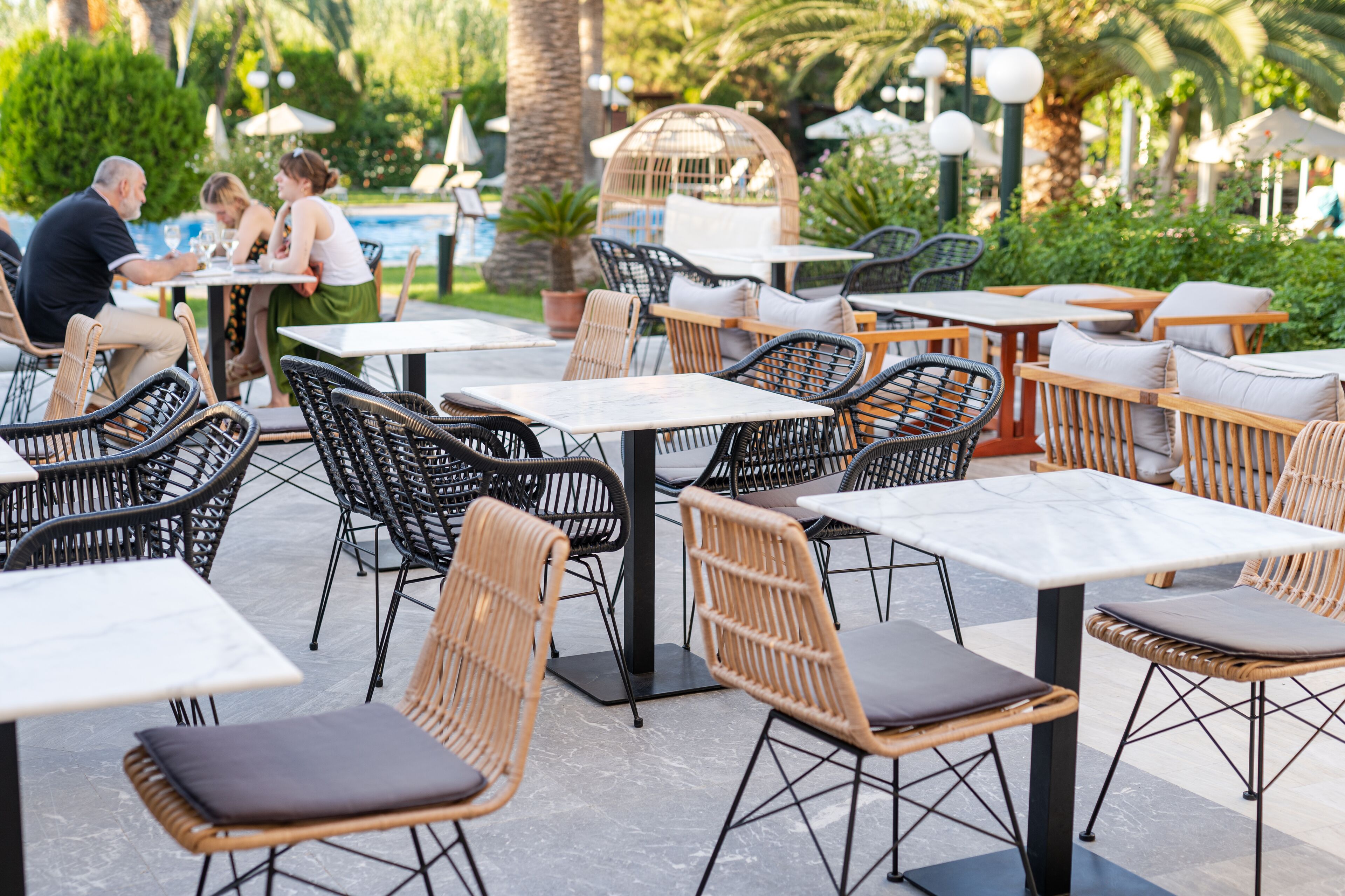 Un espace extérieur aménagé pour les repas, avec des chaises et des tables en osier, entouré de verdure et d’une piscine.