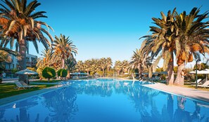 A swimming pool surrounded by palm trees and sun loungers, with a clear blue sky above. - May Beach Hotel (Rethymno)