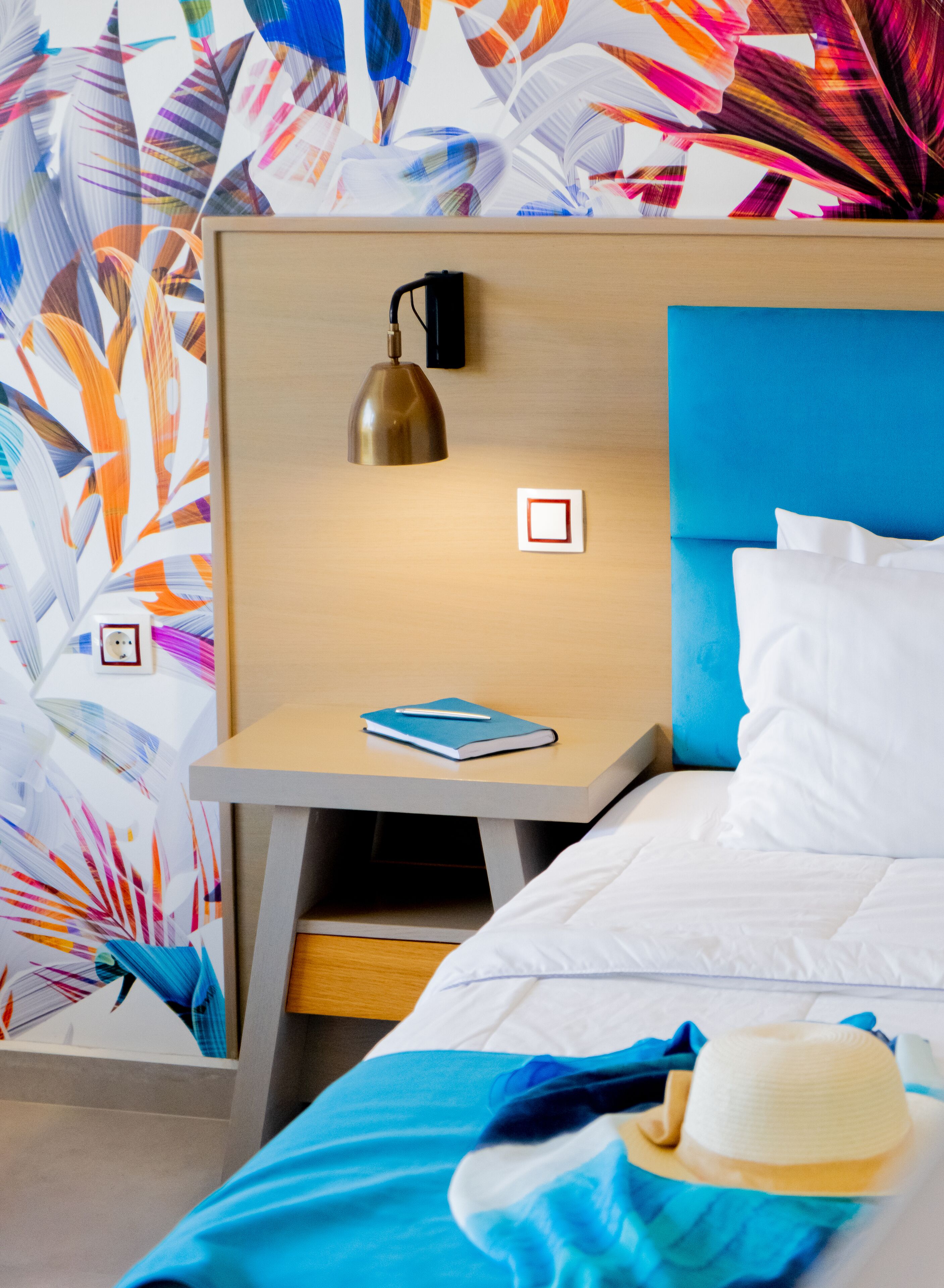 Habitación de hotel con cama, mesita de noche, lámpara y un muro con estampado de hojas de colores.