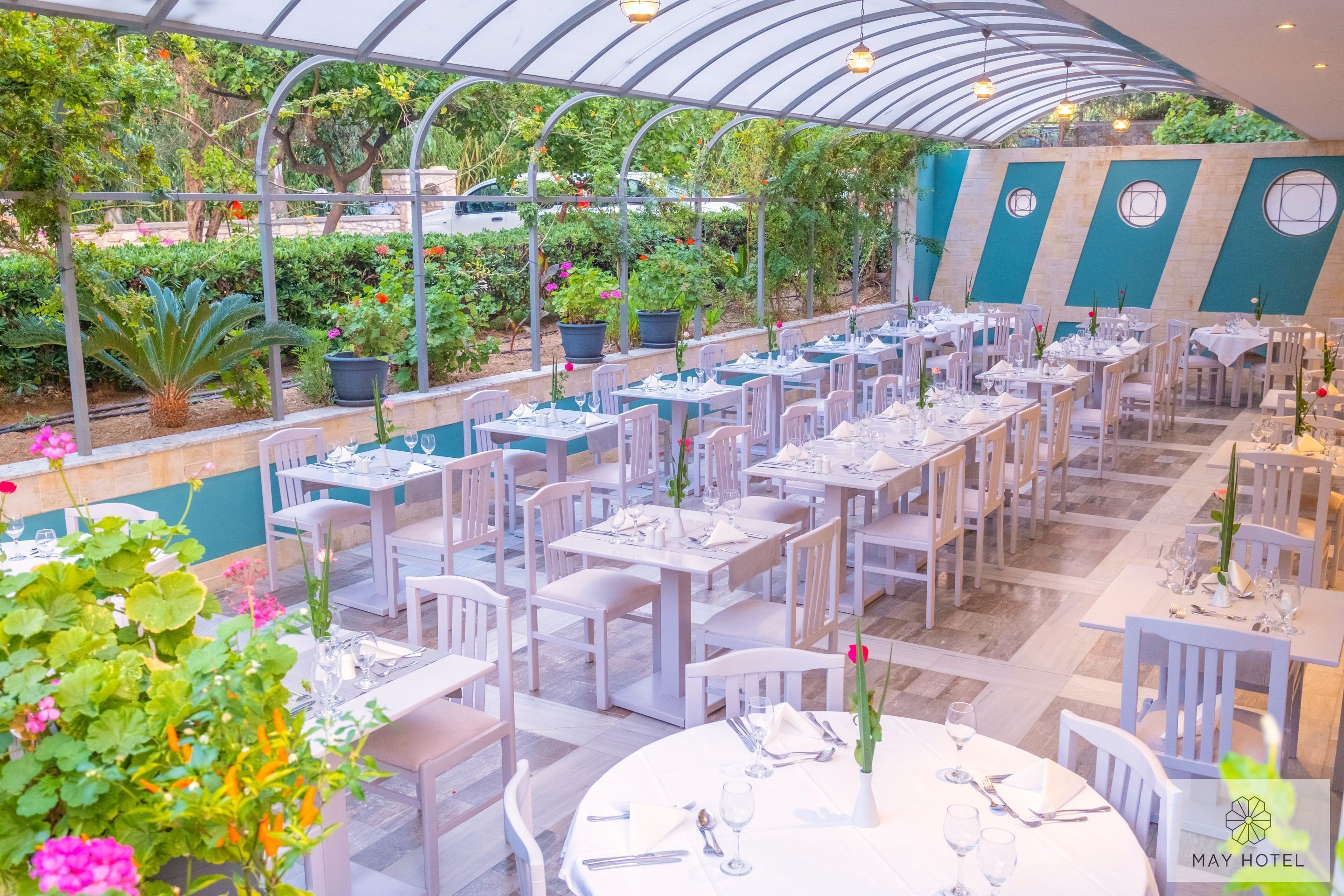 En restaurang med vita bord och stolar, omgiven av krukväxter och blommor.