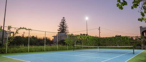 Un court de tennis en plein air, doté d’un filet, entouré d’une clôture et éclairé par deux lampadaires.