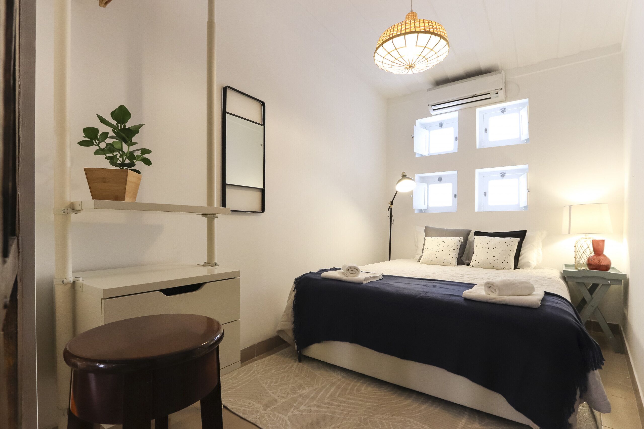 Un dormitorio con cama, mesita de noche, lámpara, planta en maceta y ventana de varios cristales.