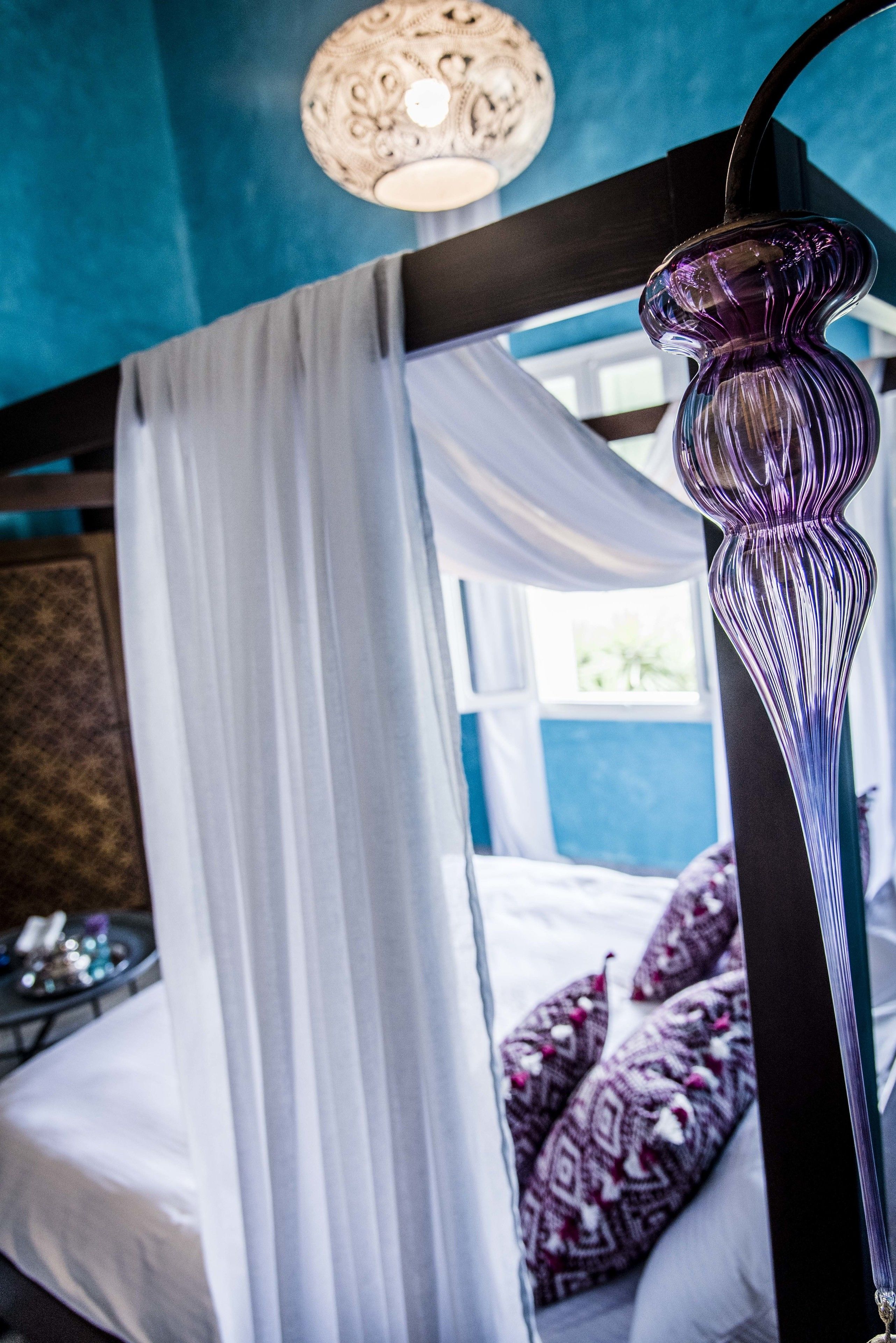 A bedroom with a bed, white curtains, a purple vase, and a ceiling light fixture.