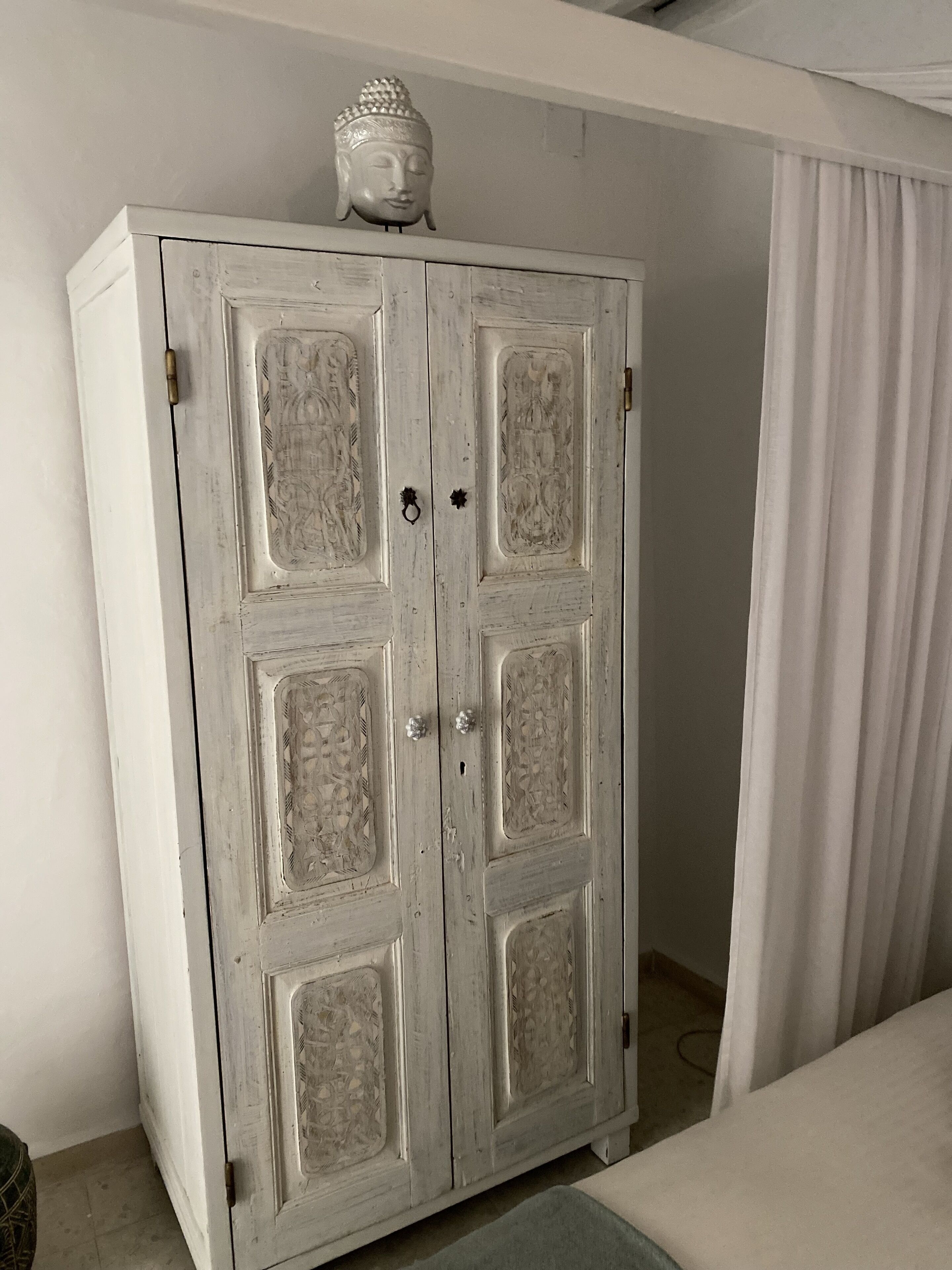 A white, vintage-style wardrobe with intricate carvings and a Buddha headpiece on top.
