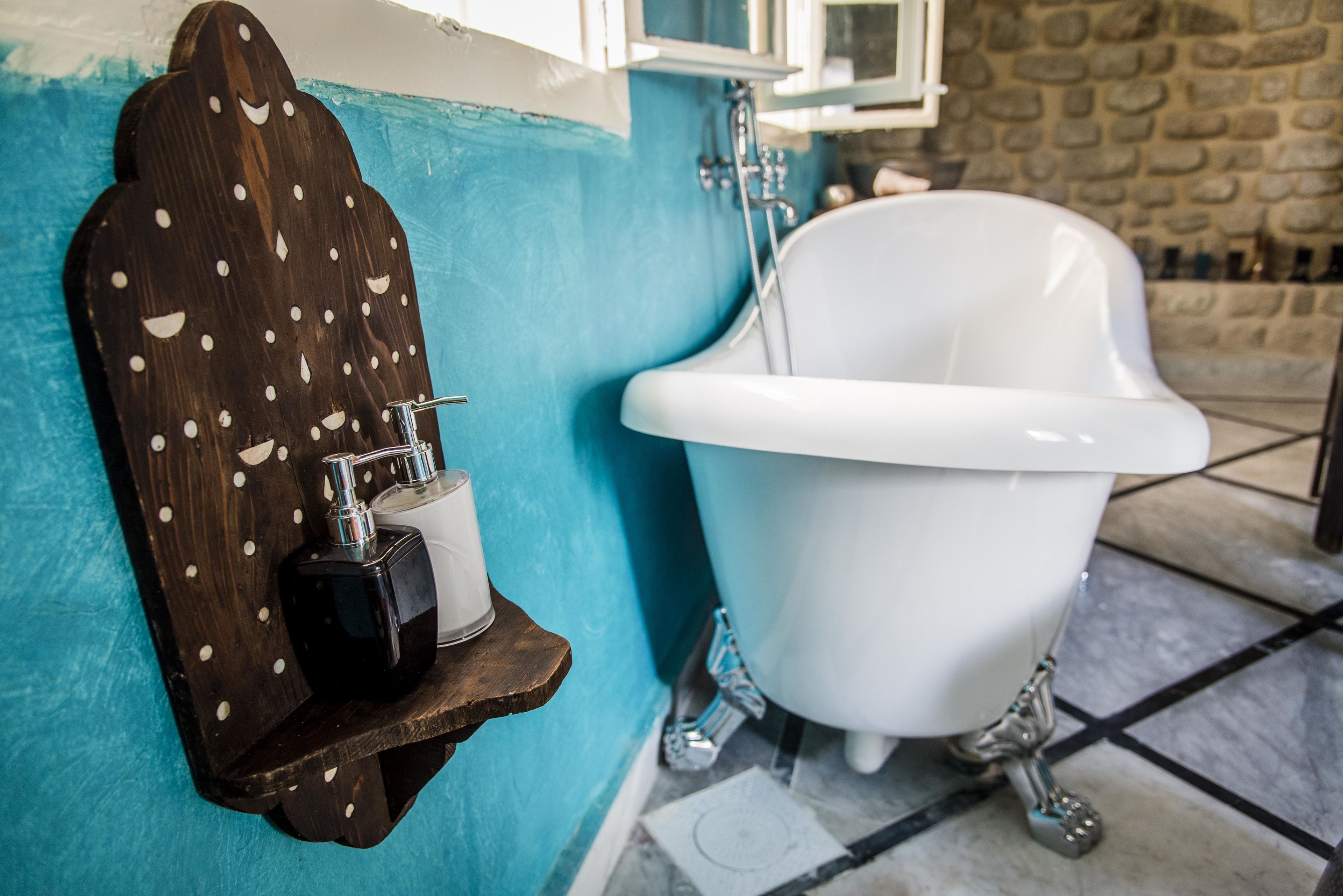 A bathroom with a white clawfoot bathtub, a wooden shelf with toiletries, and a stone wall.