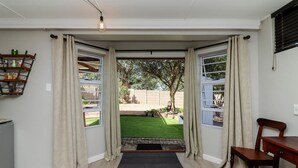A room with a view of a green lawn and trees, a wooden floor, a black mat, and a chair. - Arbour1 (George)