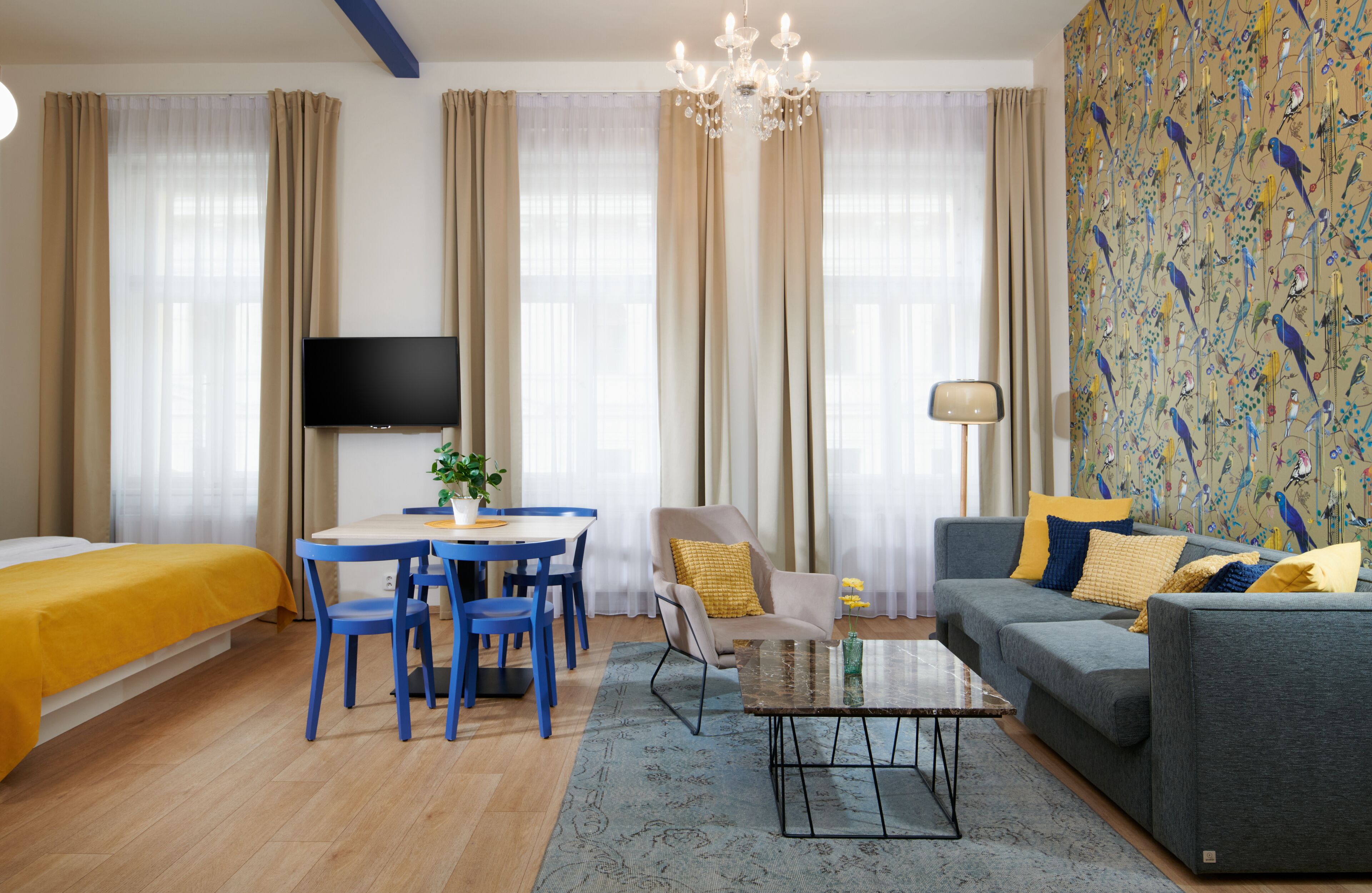 A modern hotel room with a bed, a sofa, a dining table with chairs, a TV, and a patterned wall.