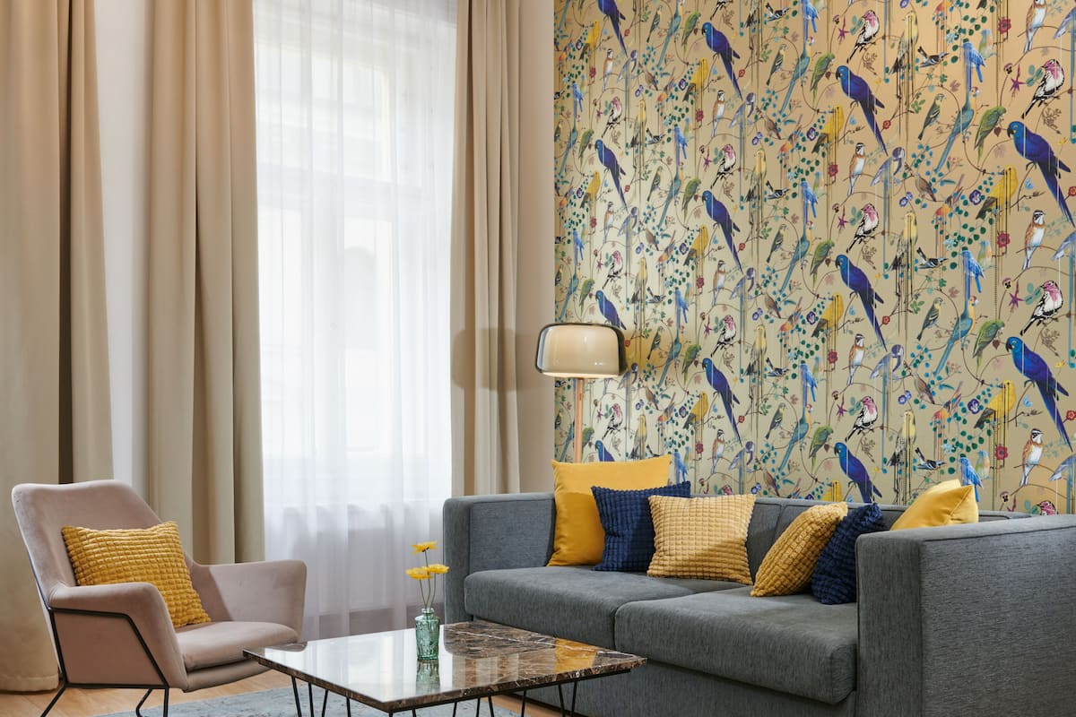 A living room with a grey sofa, a matching armchair, and a glass coffee table. A patterned wallpaper with birds and flowers adorns the wall.