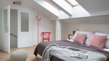 Quarto com claraboia, cama com almofadas, uma cadeira vermelha e um banquinho redondo.