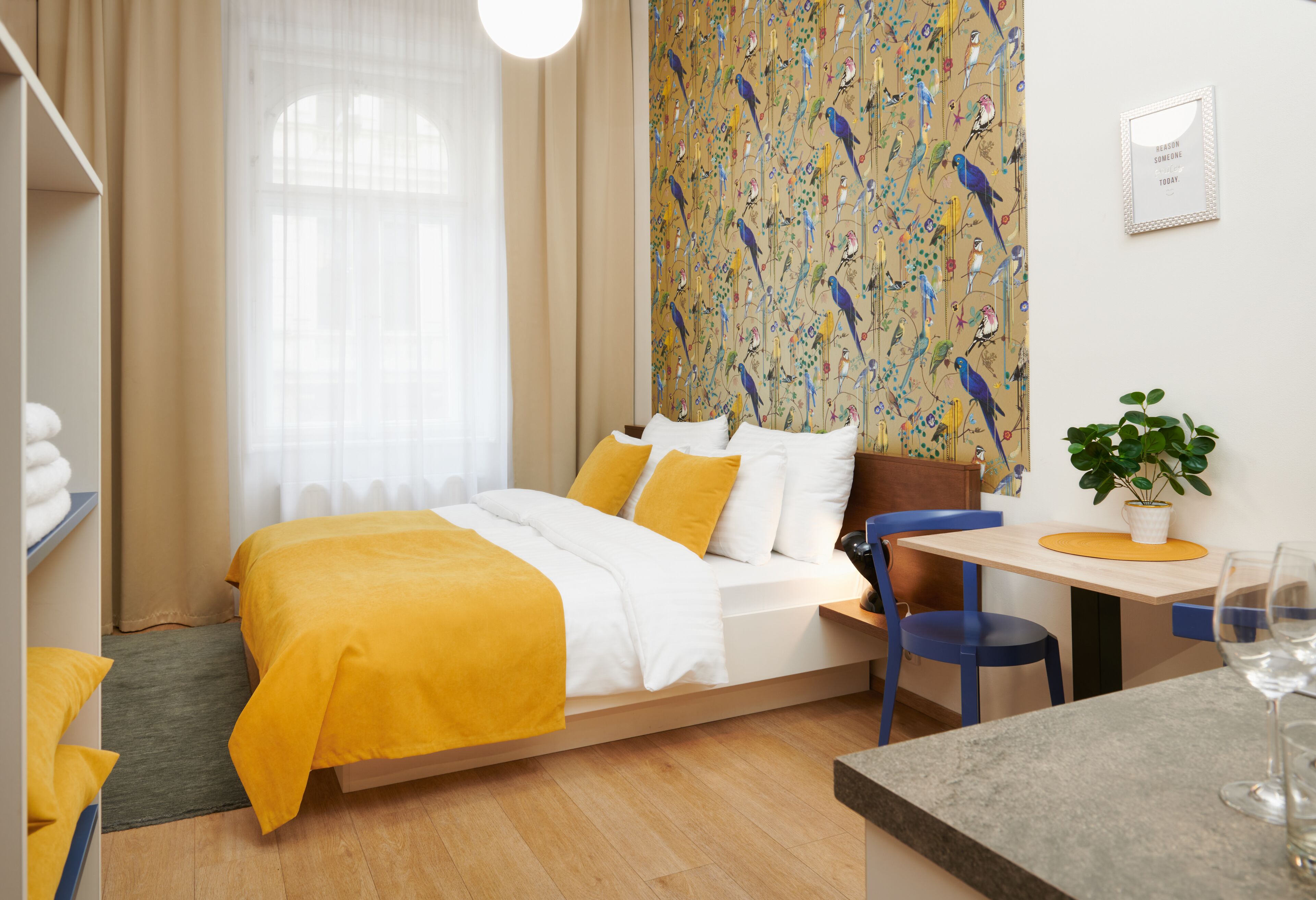 A modern hotel room with a bed, a desk, a chair, a plant, and a window with curtains.