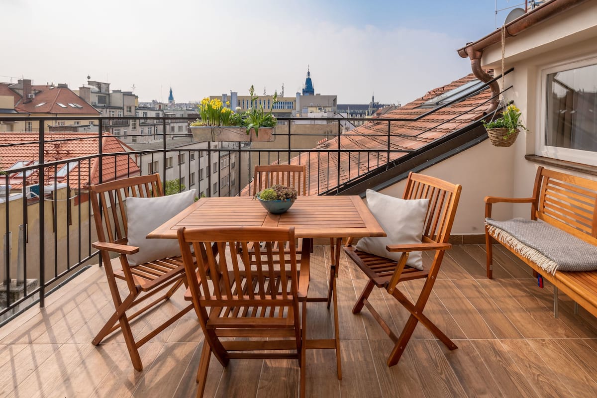A wooden table and chairs on a balcony with a city view, potted plants, and a bench.