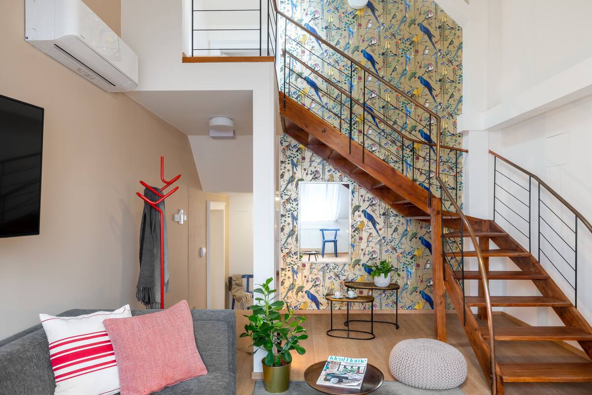 A modern living room with a grey sofa, a coffee table, and a staircase with a bird-patterned wallpaper.