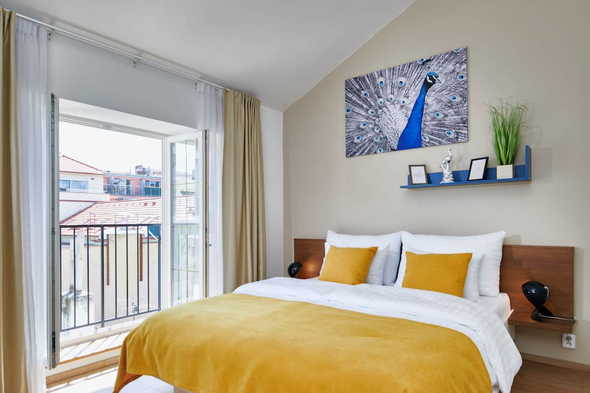 A hotel room with a large bed, a balcony with a view of buildings, a wall-mounted shelf with decorative items, and a painting of a peacock.