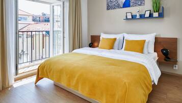 A hotel room with a large bed, a balcony with a view of buildings, a wall-mounted shelf with decorative items, and a painting of a peacock.