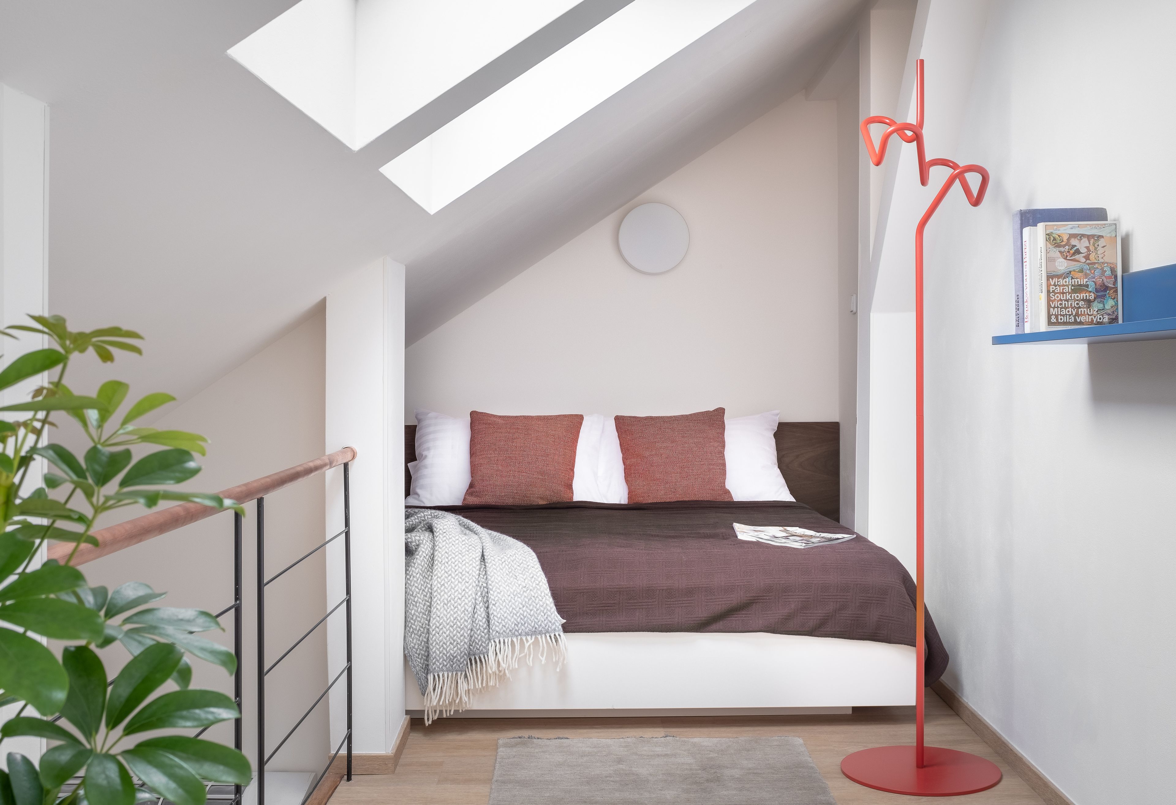 A bedroom with a sloped ceiling, a bed with a brown blanket, a red floor lamp, and a small bookshelf.