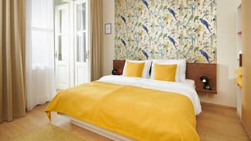 Une chambre d’hôtel avec un grand lit, une couvre-lit jaune, une tête de lit en bois et un mur orné d’un motif représentant des oiseaux.
