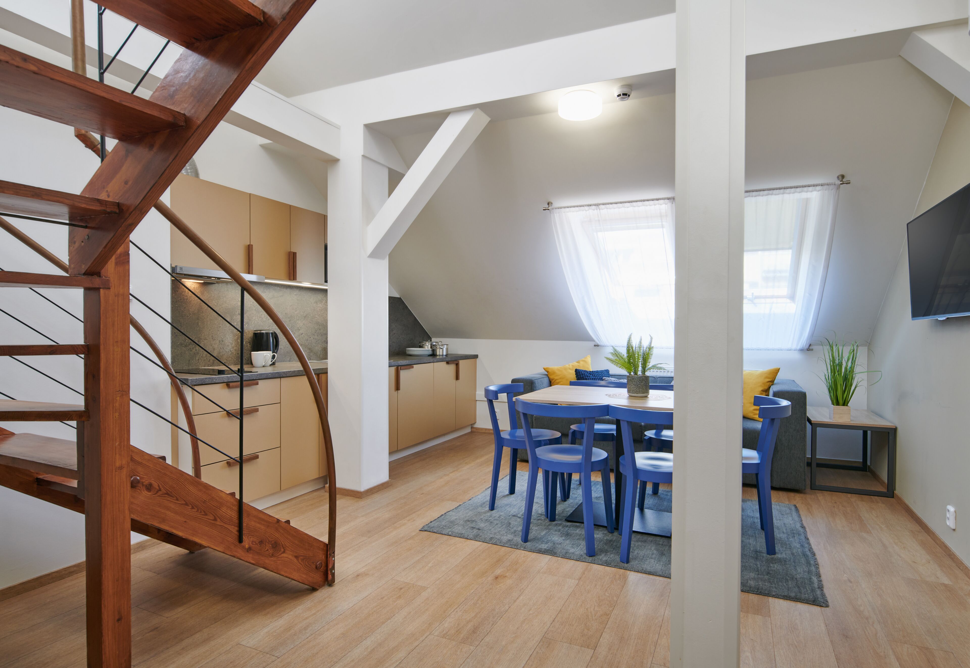 Un appartement moderne de style « loft », doté d’un escalier en bois, d’une cuisine, d’un coin repas avec des chaises bleues et d’un espace de vie avec un téléviseur.