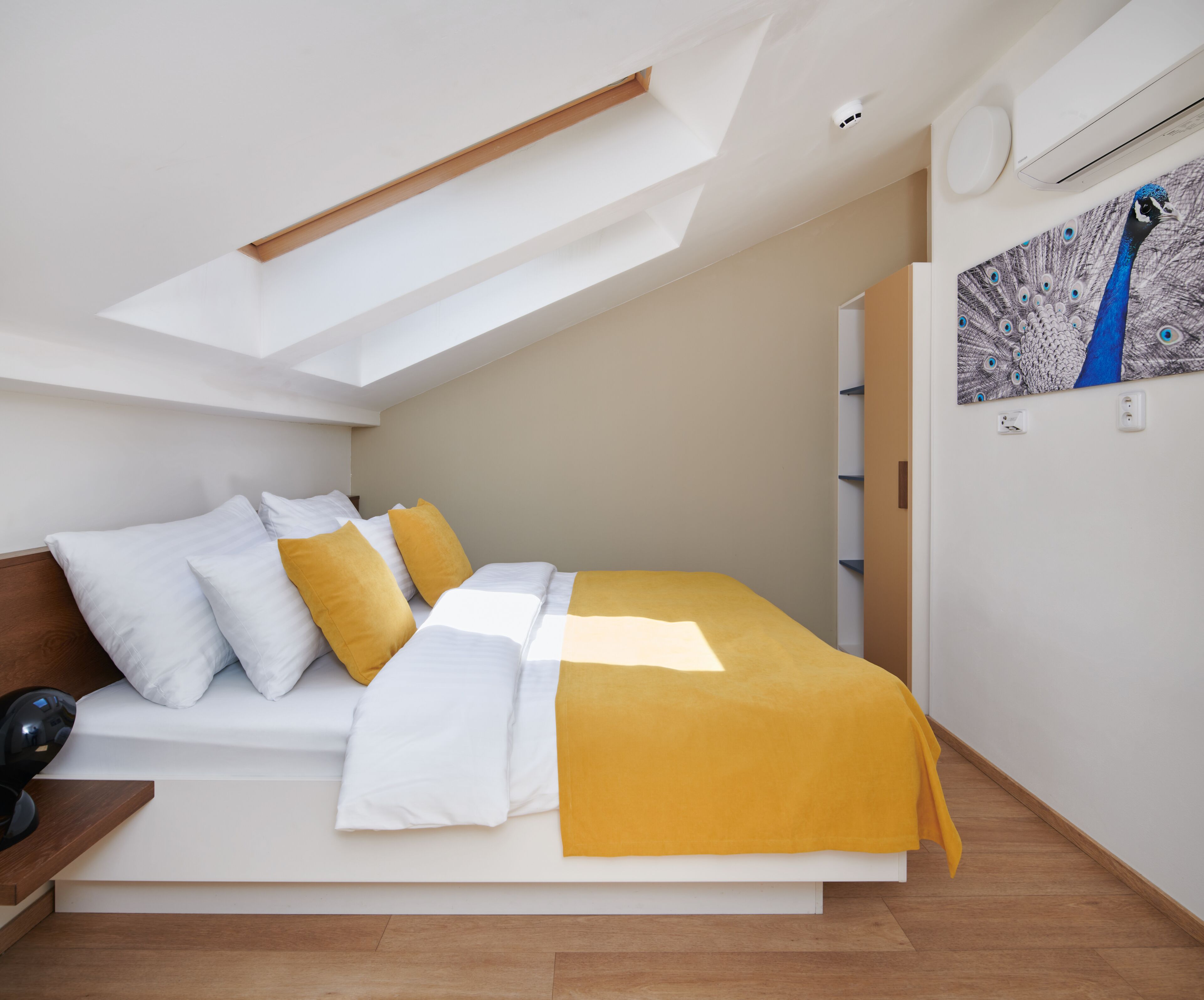 Ein modernes Schlafzimmer mit einem Oberlicht, einem Bett mit weißer und gelber Bettwäsche, Holzboden und einem Wandbild mit einem Pfau.