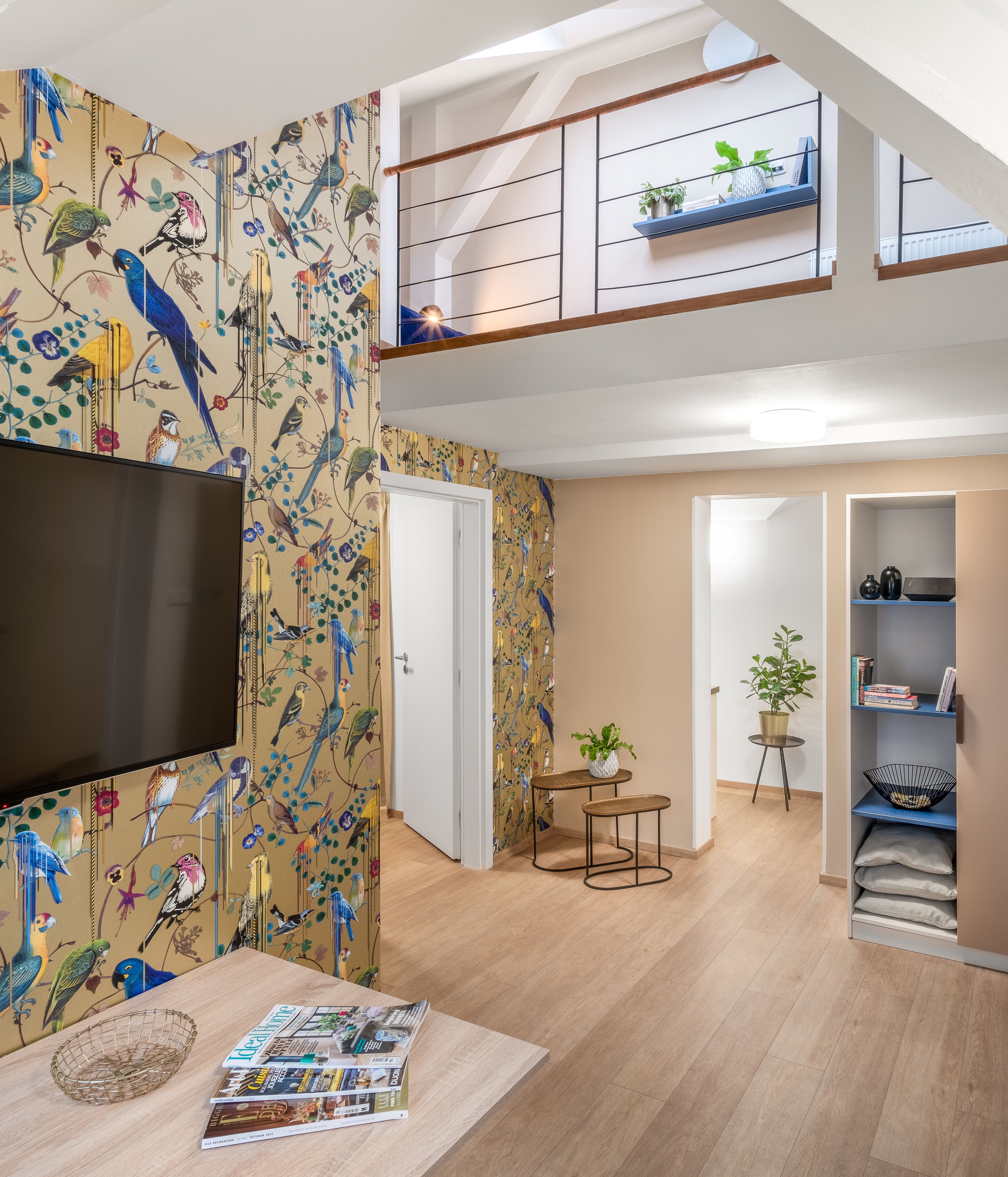 A modern apartment with a large TV, a dining area with magazines, a staircase, and a balcony with plants.
