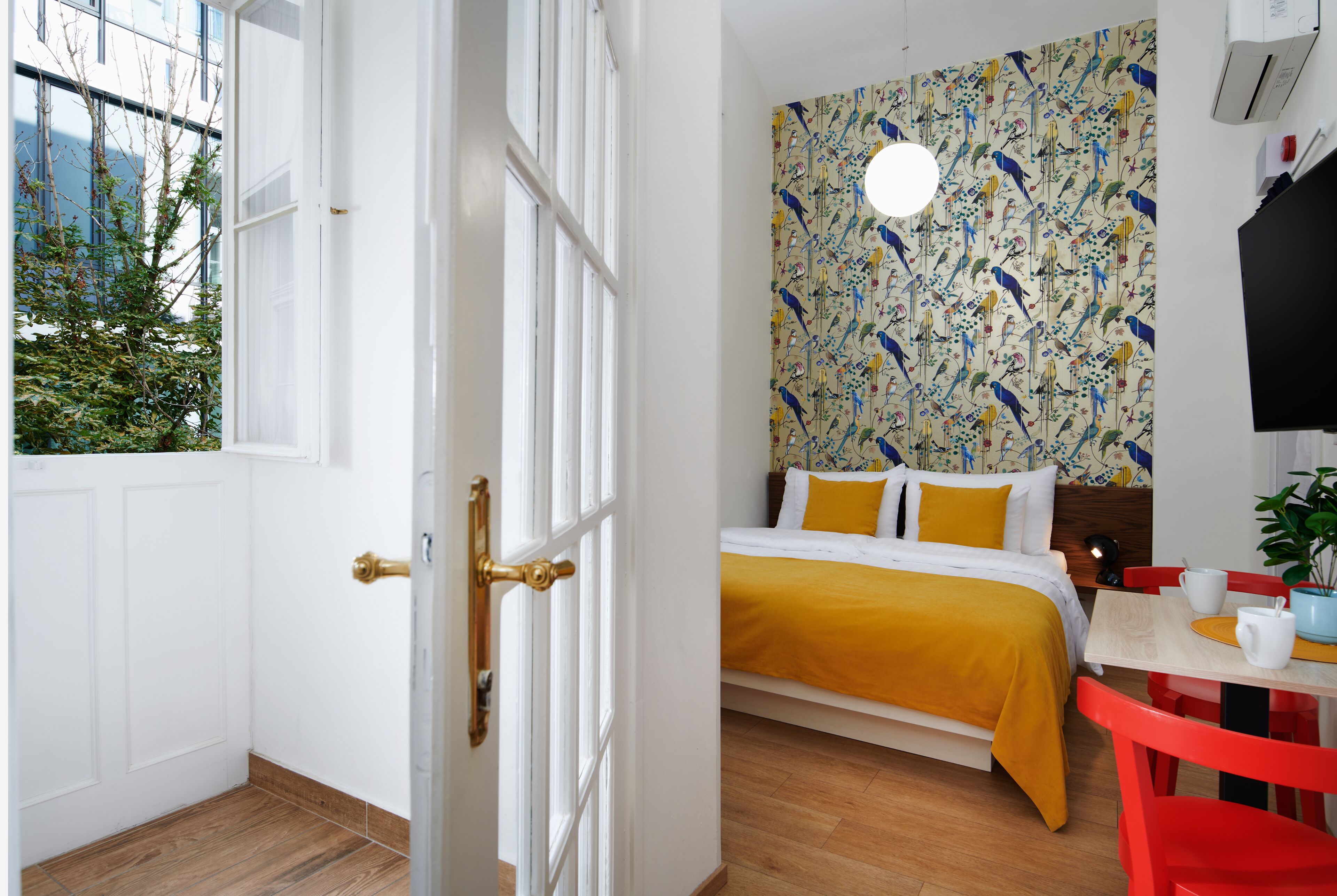 A bedroom with a bed, a window, a wall with a bird pattern, a table with a plant, and a red chair.