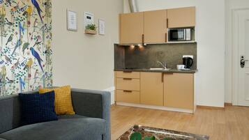 A modern living room with a grey sofa, a coffee table, and a colorful wall mural. Adjacent is a compact kitchen with a microwave, coffee maker, and wooden cabinets.