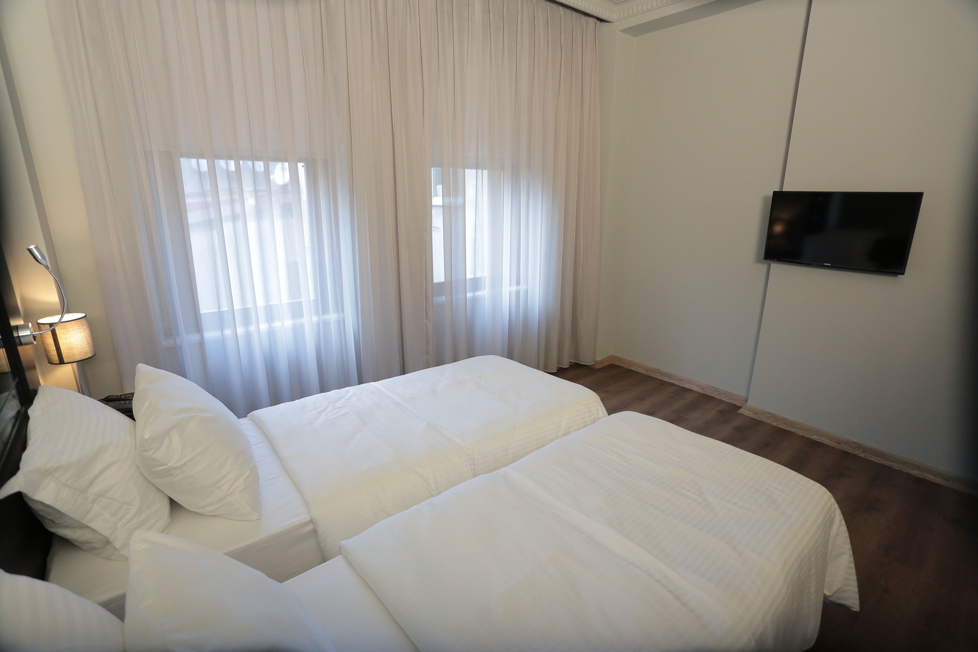 A hotel room with two beds, a TV mounted on the wall, and a window with sheer curtains.