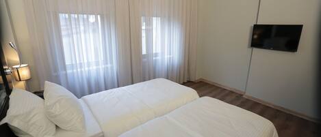 A hotel room with two beds, a TV mounted on the wall, and a window with sheer curtains.