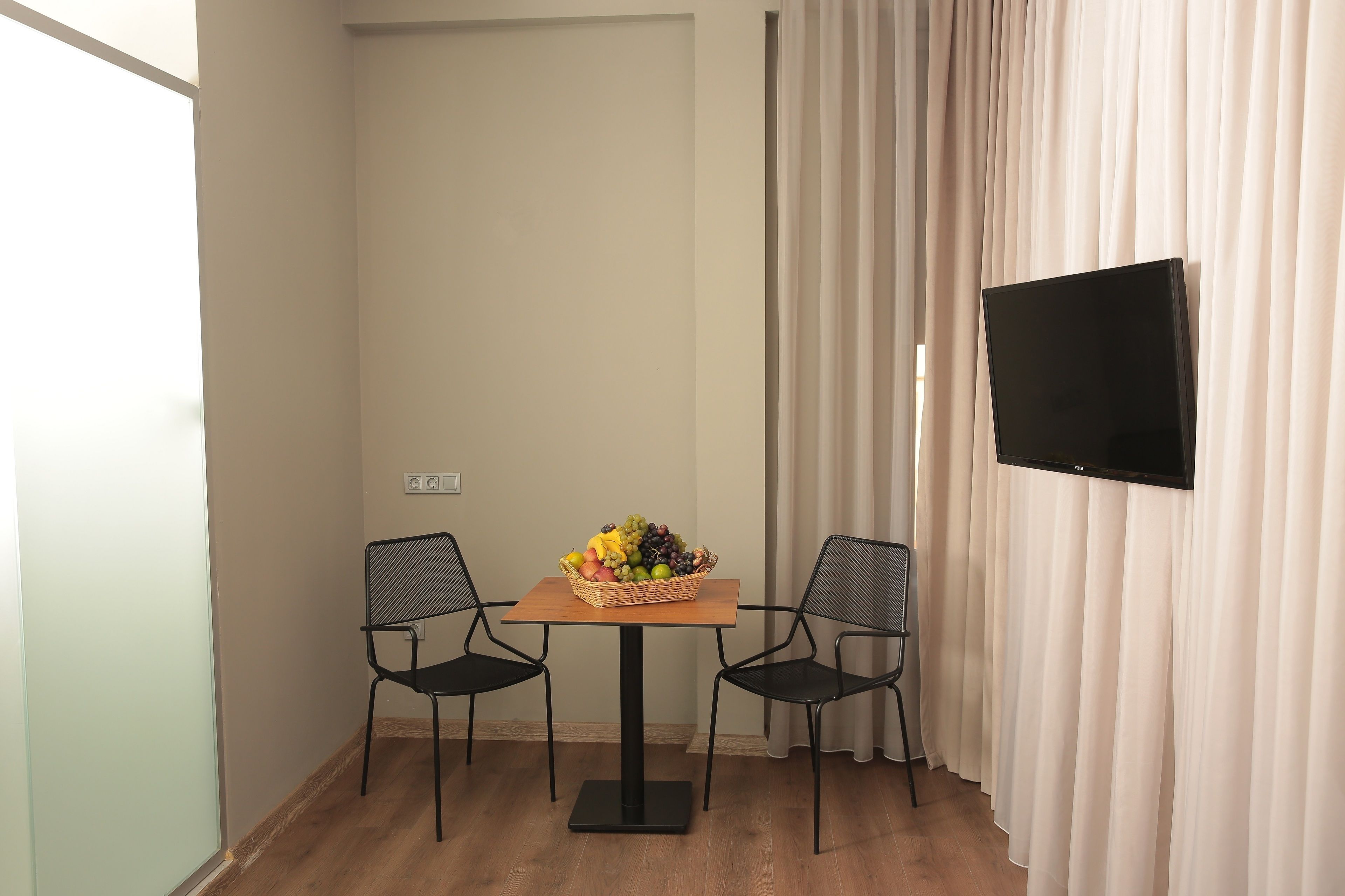 Una habitación pequeña con una mesa de madera, dos sillas, una cesta de frutas, un televisor de pantalla plana y un ventanal con cortinas.