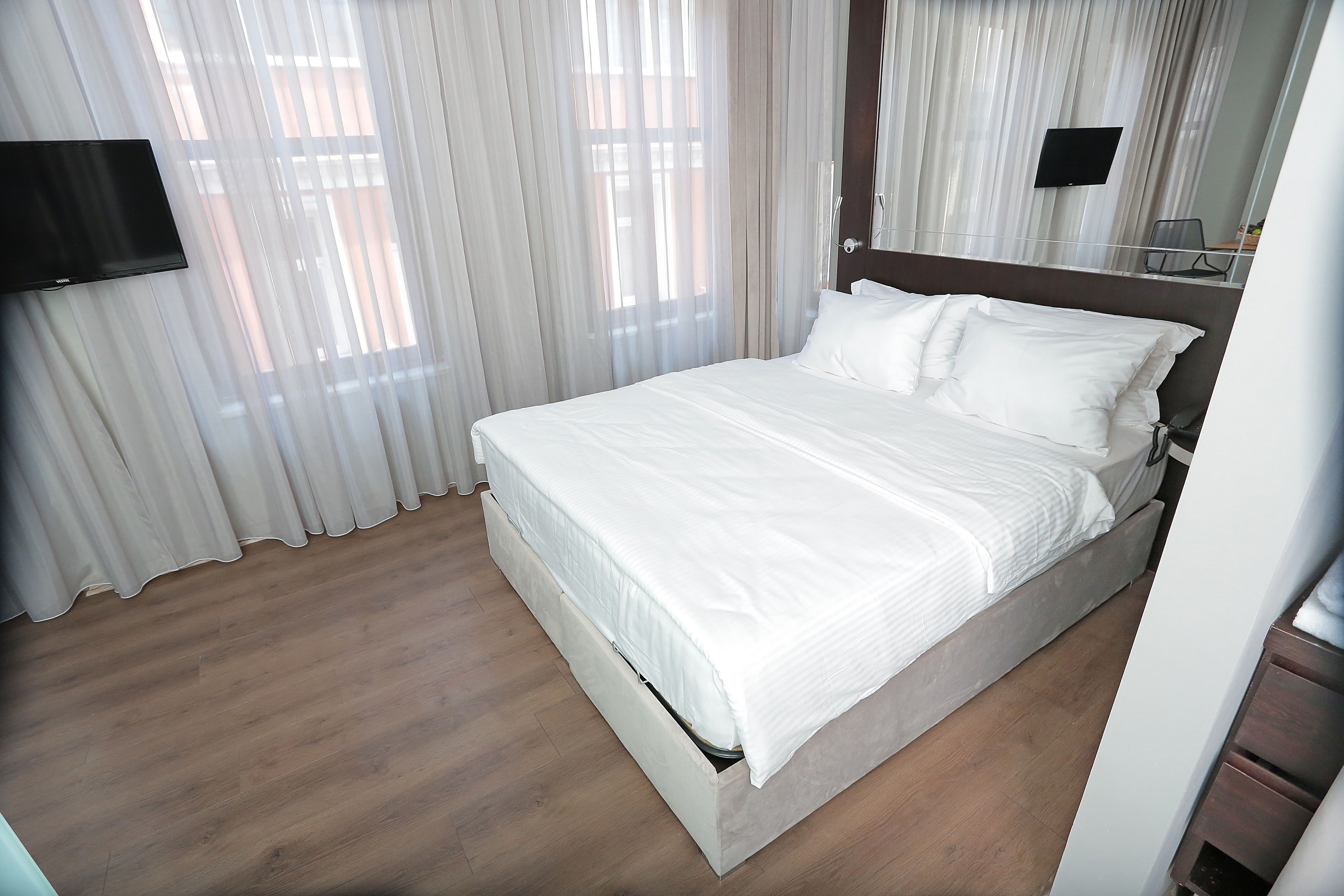A hotel room with a bed, curtains, a flat-screen TV, and a wooden floor.