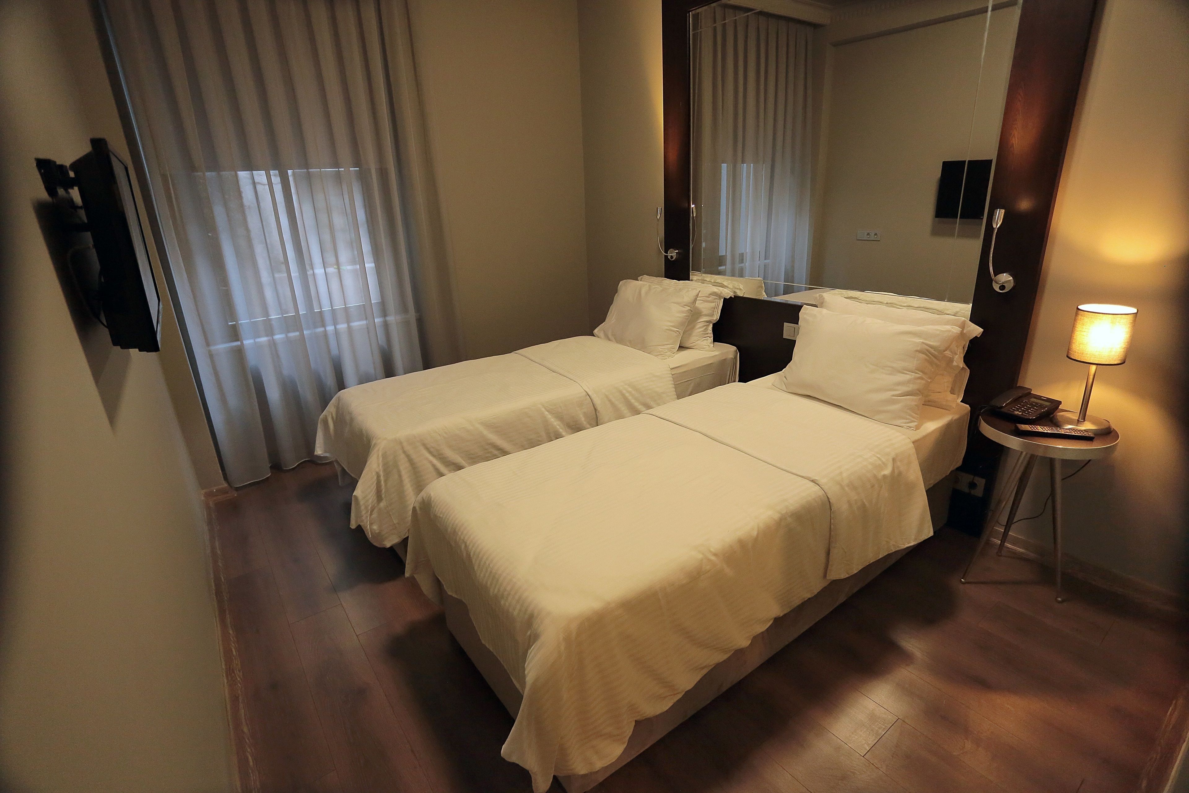 Habitación de hotel con dos camas, una mesita de noche con lámpara, un espejo y una ventana con cortinas.