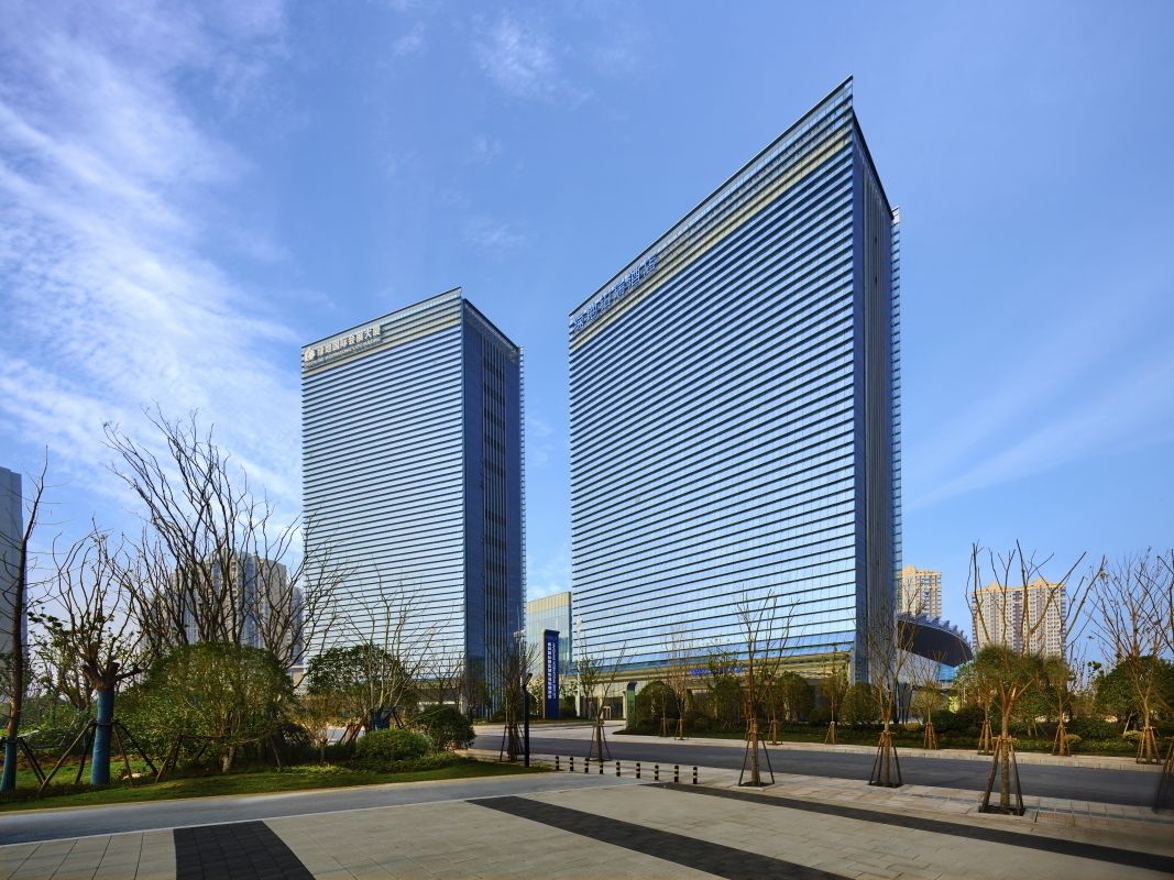 Dos edificios modernos de gran altura con fachadas de vidrio reflectante, rodeados de un área ajardinada con árboles y un camino pavimentado.