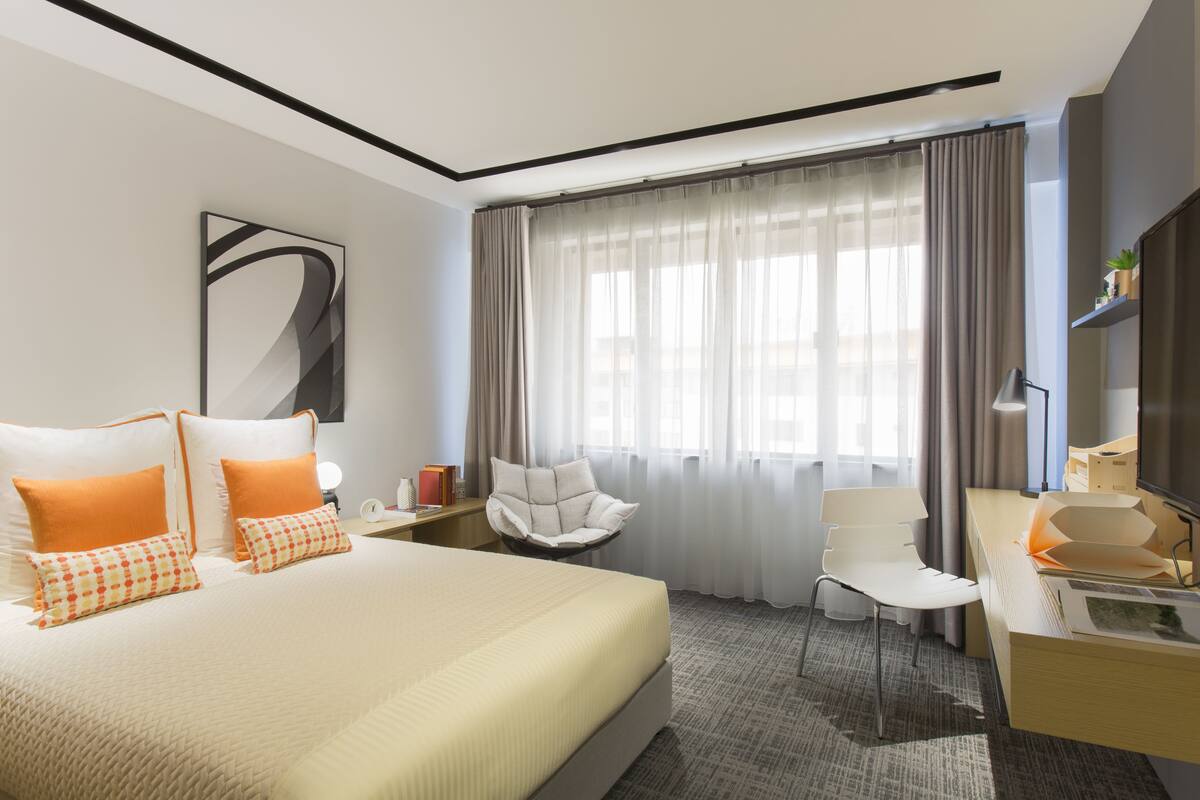 Una habitación de hotel moderna con una cama grande, dos sillas blancas, un escritorio con lámpara y un amplio ventanal con cortinas translúcidas.