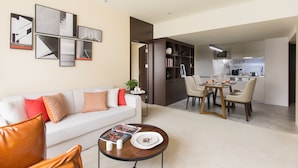 Un salon moderne avec un canapé blanc, une table basse et un coin repas comprenant une table et des chaises. On aperçoit une cuisine à l’arrière-plan, avec des placards et des appareils électroménagers.