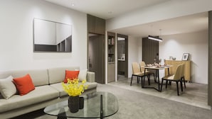 Un salon moderne avec une table basse en verre, un canapé avec des coussins orange, un coin repas avec des chaises et une table, et une cuisine en arrière-plan.