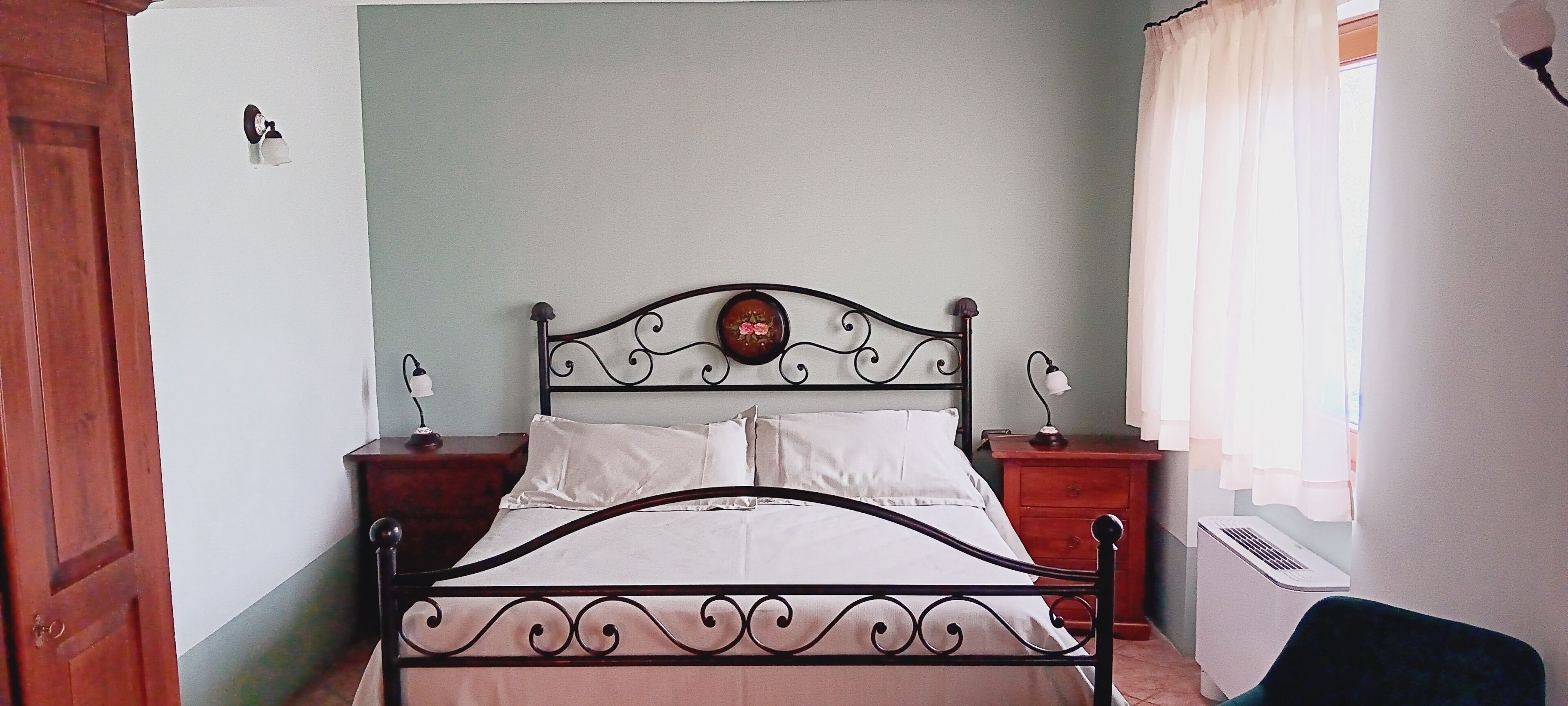 A bed with a metal frame and a wooden headboard, two bedside tables with lamps, a wall clock, and a window with curtains.