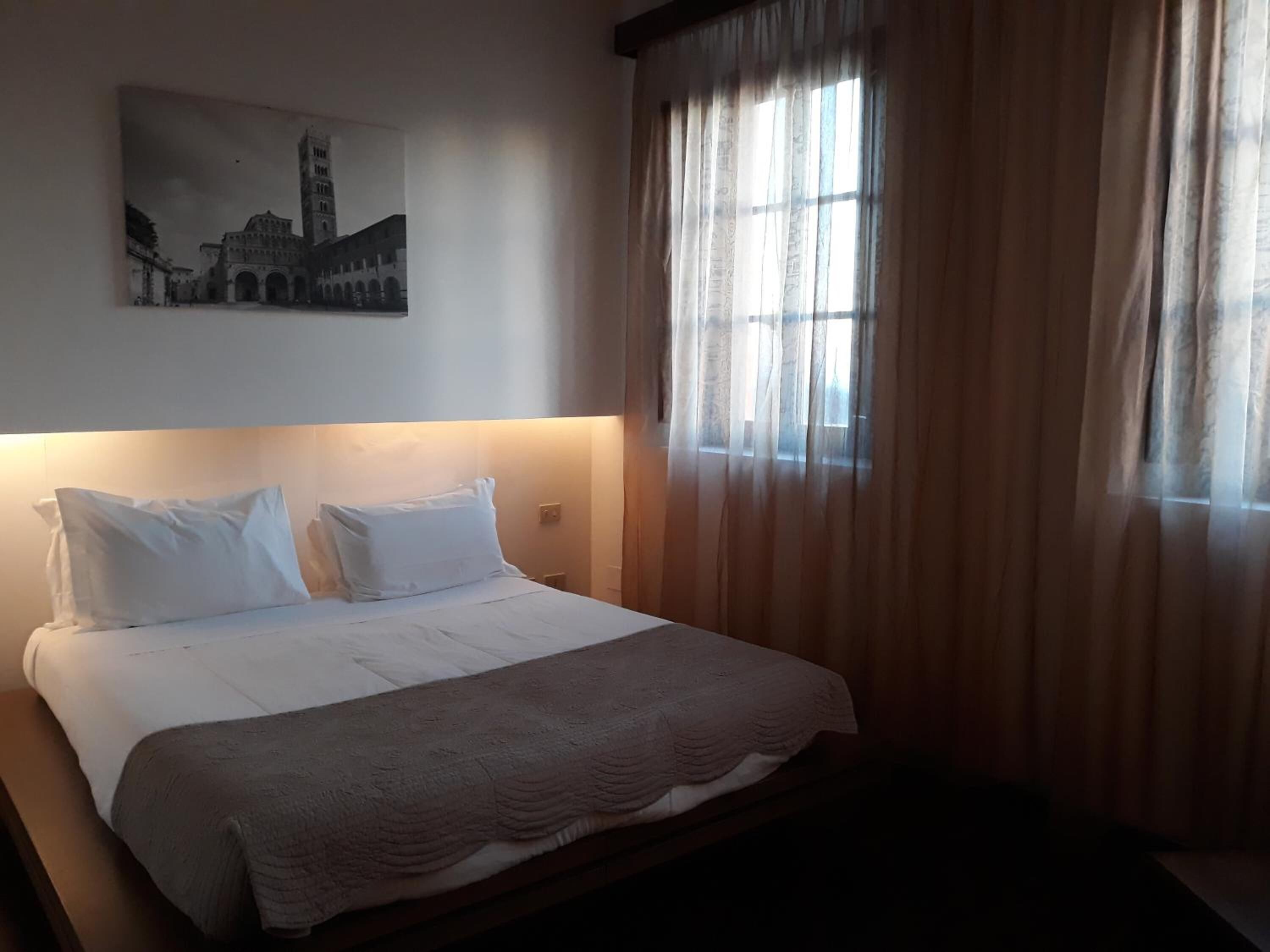 Een hotelkamer met een net opgemaakt bed, een groot raam met dunne gordijnen en een lijst met een afbeelding van een gebouw aan de muur.