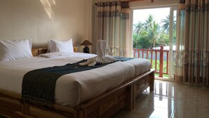 A hotel room with a large bed, a wooden headboard, a nightstand with a lamp, and a view of palm trees through the window.