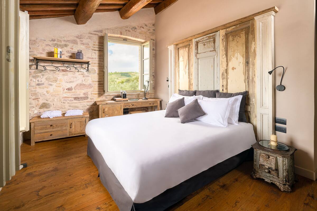 A bedroom with a large bed, wooden headboard, stone wall, wooden floor, and a rustic dresser.