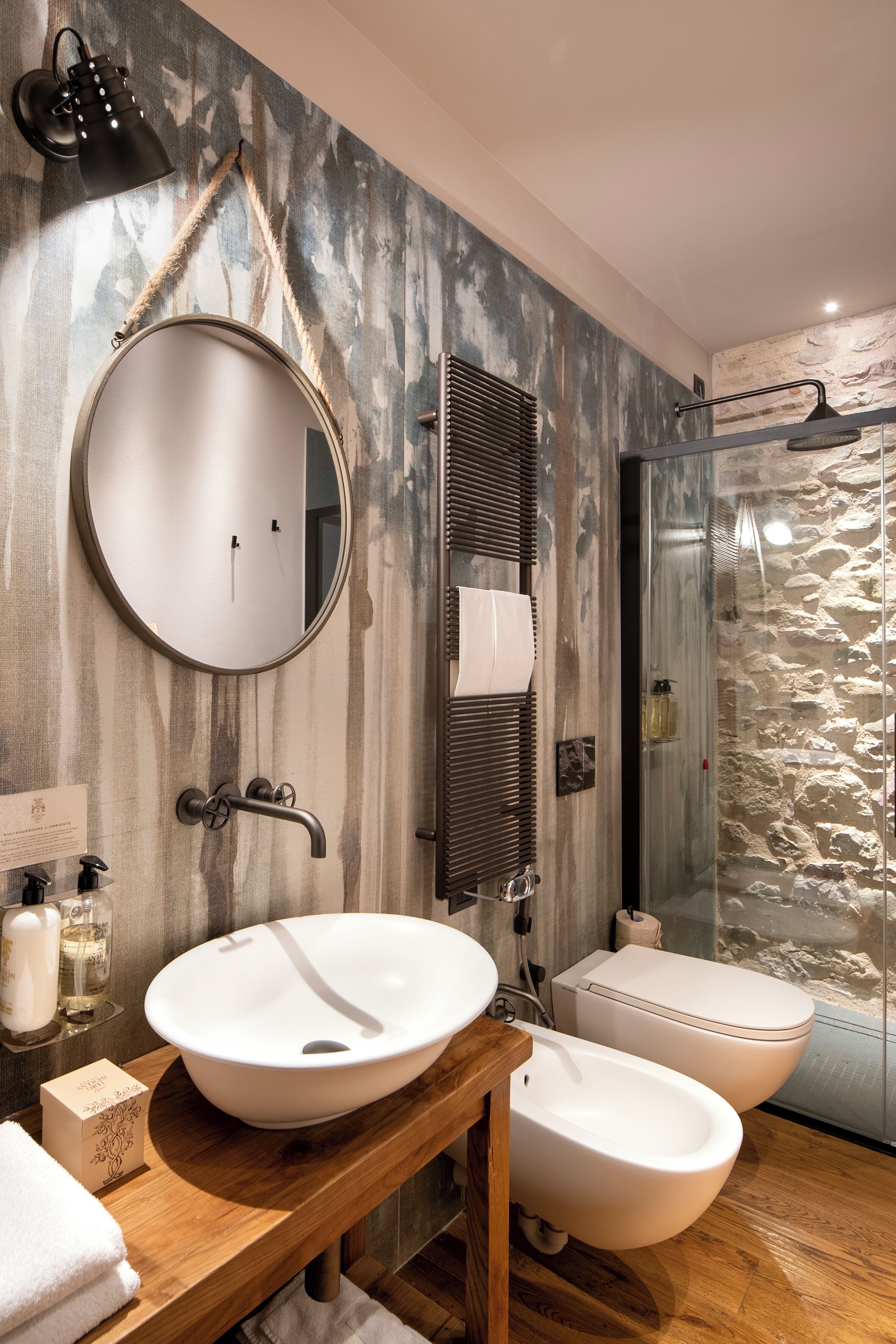 Une salle de bain avec un meuble-lavabo en bois, un miroir rond, un lavabo blanc, deux bidet, un porte-serviettes et une douche à parois de verre.