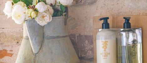 A bathroom with a stone wall, a vase with white flowers, two bottles of Antichi Orti body wash, and a folded white towel on a wooden surface.