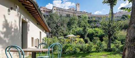 A courtyard with a table and chairs, surrounded by greenery and a building with arches in the background.