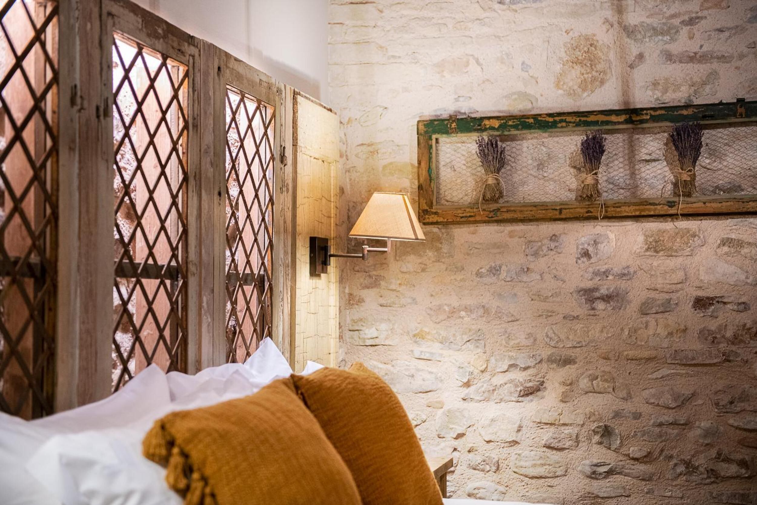 A room with a stone wall, a bed with white and mustard-colored bedding, and a wall-mounted lamp.