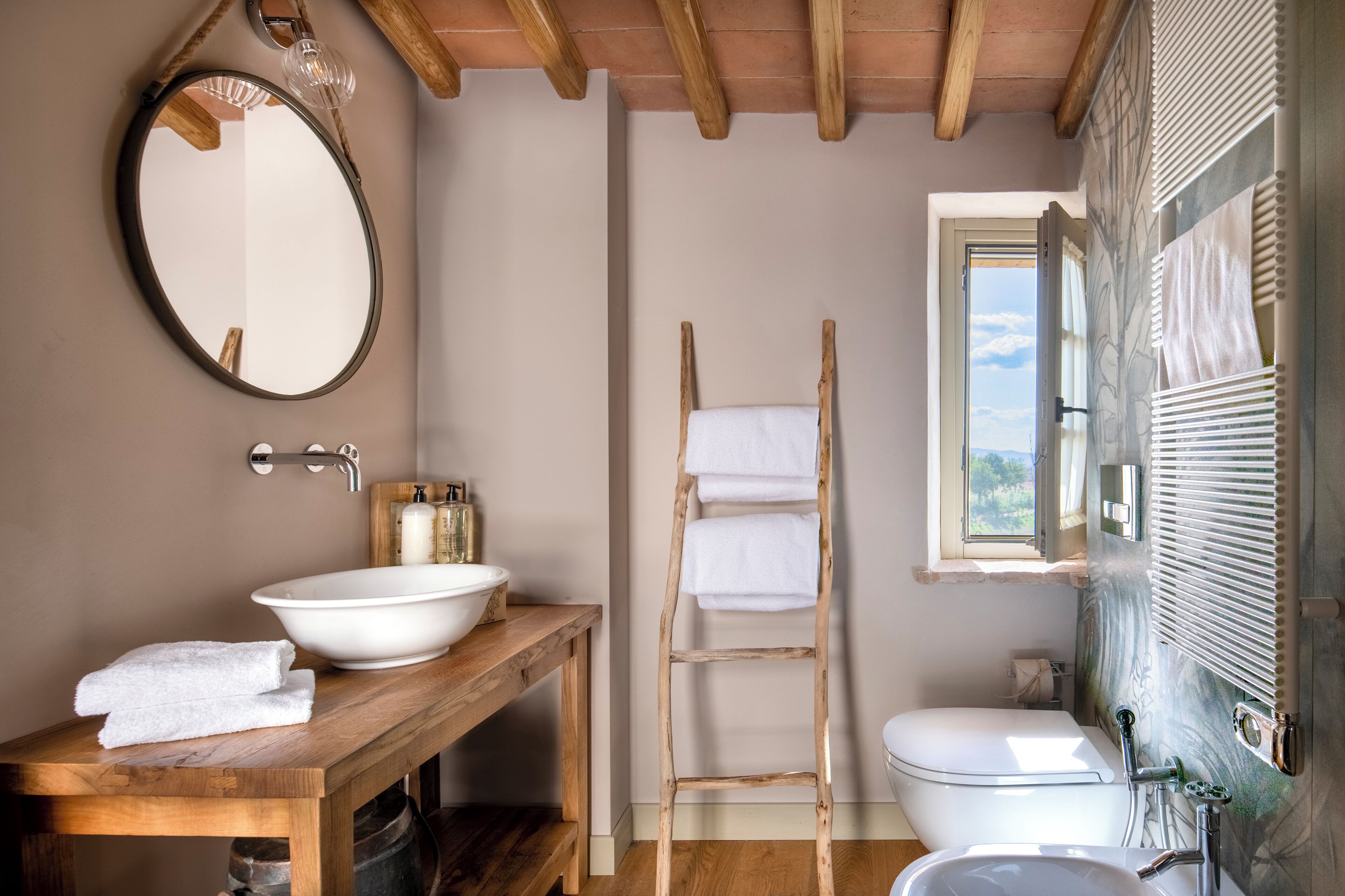 A bathroom with a wooden vanity, a round mirror, a white sink, a wooden ladder, and a toilet.